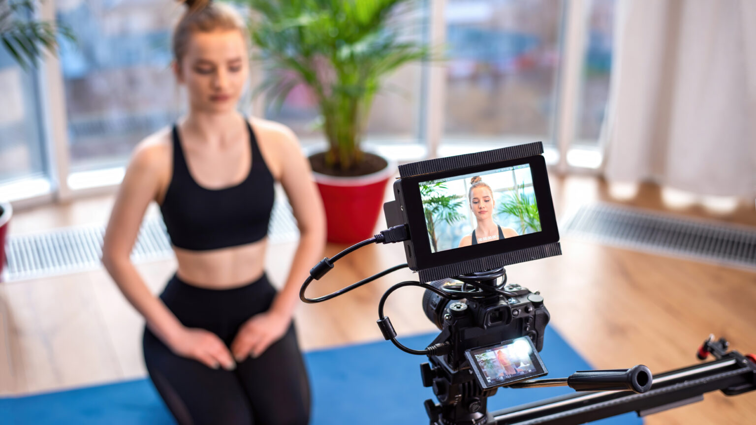Professional camera with external display recording a young blonde woman in sportswear while meditating on a yoga mat. Panoramic windows